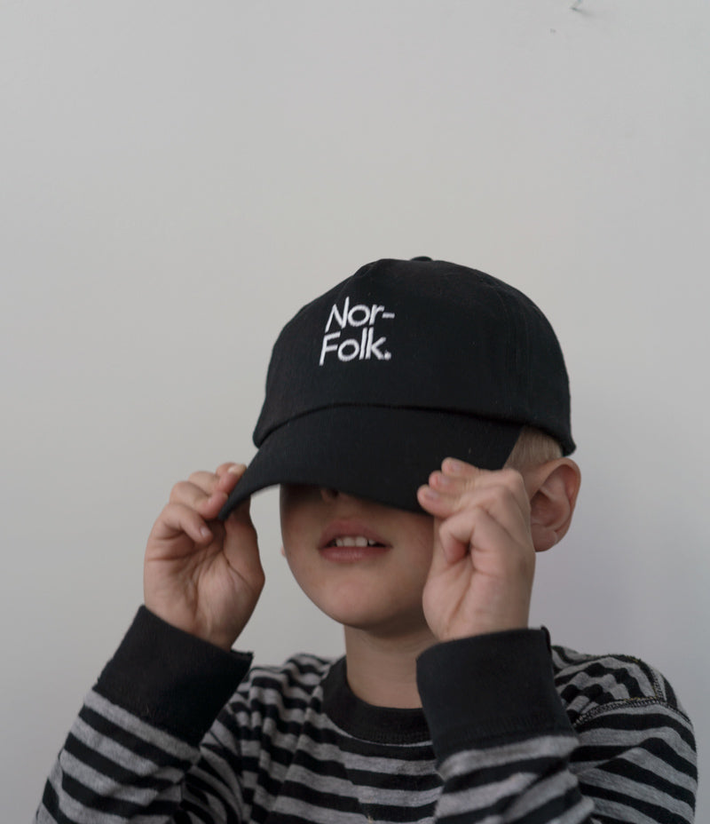 Image shows a child wearing a black baseball cap pulled over their eyes. On the cap  is the 'Nor-folk' logo in white. Nor–Folk Basics Kids Cap - Nor–Folk