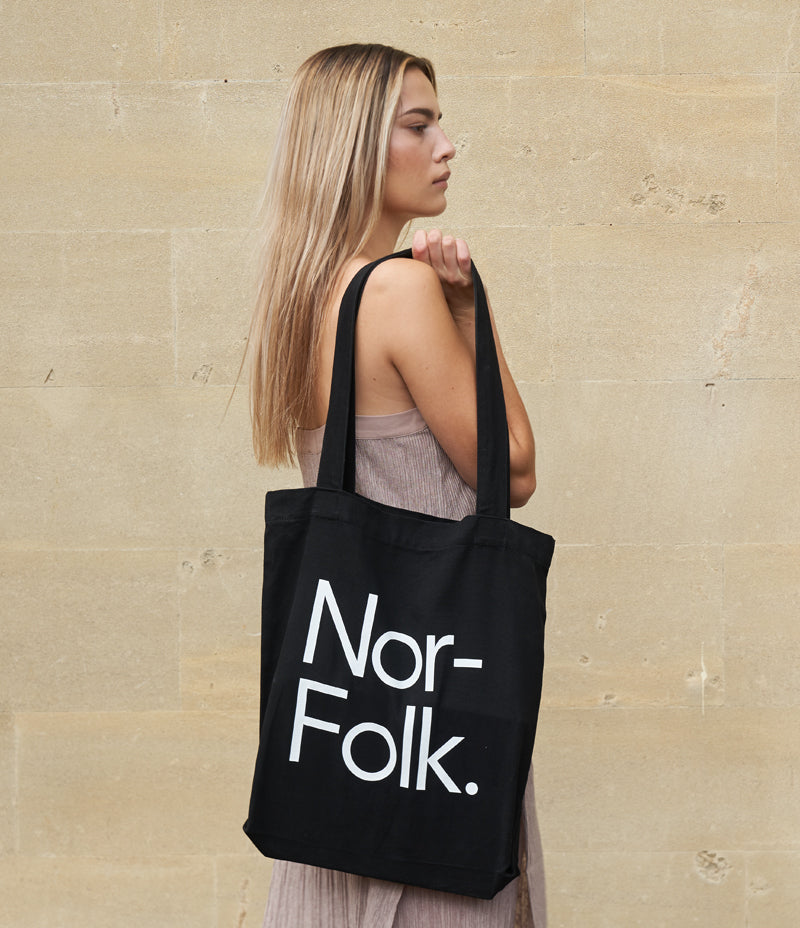 Image shows an adult female standing infant of a wall. Over her right shoulder is a black tote bag with the 'Nor-Folk' logo in white. Nor–Folk Basics Tote Bag - Nor–Folk