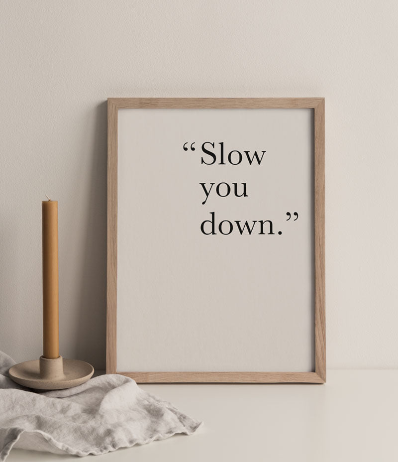 Image shows a print in a wooden frame. The background on the print is cream with the words 'Slow You Down' in black text. To the left of the print  is an orange candle in a ceramic holder and a linen cloth.  Print ~ Coming soon. - Nor–Folk